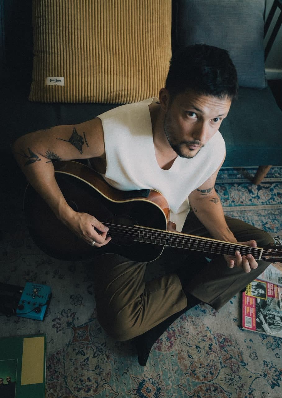 Tyan seated on a rug with an acoustic guitar.